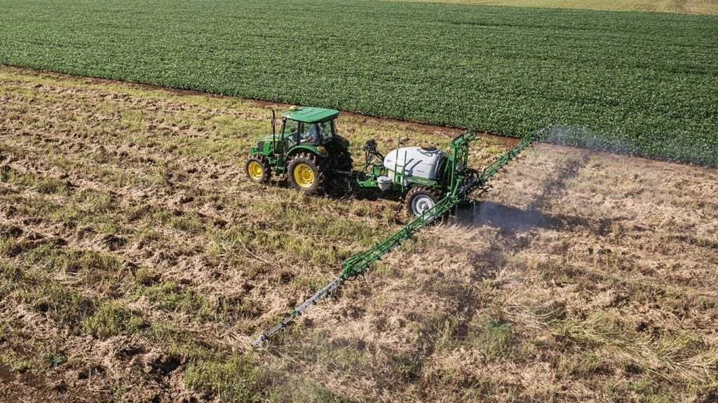 A imagem, capturada de um ângulo elevado, mostra uma operação agrícola em pleno andamento. Um trator verde da marca John Deer