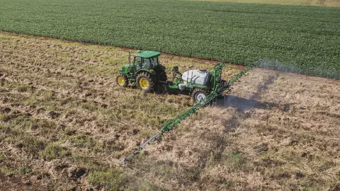A imagem, capturada de um ângulo elevado, mostra uma operação agrícola em pleno andamento. Um trator verde da marca John Deer