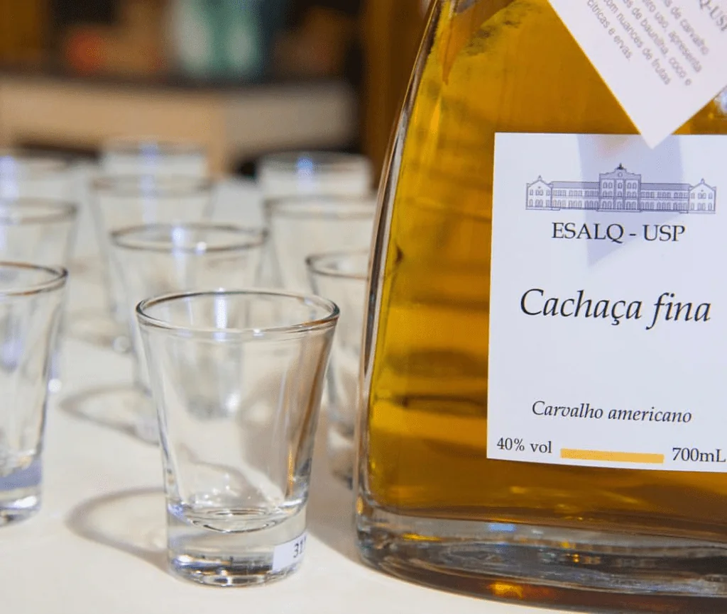 primeiro plano uma garrafa de ‘Cachaça fina’ de cor dourada, indicando envelhecimento em barris, ao lado de