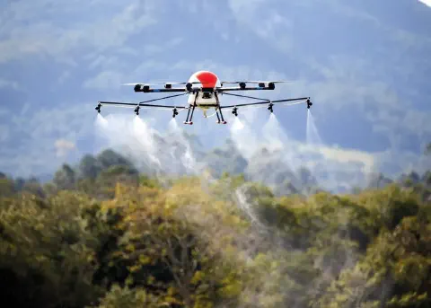 drone agrícola de grande porte em pleno voo, realizando a pulverização sobre uma área de vegetação densa. O
