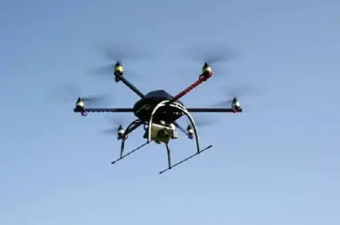 drone do tipo hexacóptero, com seis hélices em movimento, voando contra um céu azul claro e sem nuvens. O e