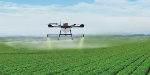 drone agrícola de grande porte em pleno voo sobre uma extensa lavoura verde, com as culturas plantadas em f