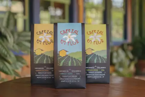 três embalagens de café da marca 'Cafezal em Flor', posicionadas verticalmente sobre uma superfície de madeira