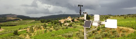 uma estação meteorológica moderna instalada em primeiro plano, contrastando com uma paisagem rural europeia ao