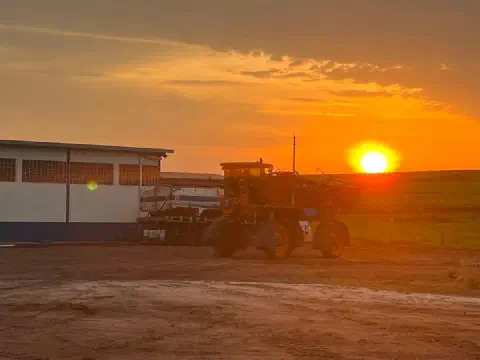 Pulverizador autopropelido estacionado ao lado de um barracão, com o sol se pondo no horizonte.
