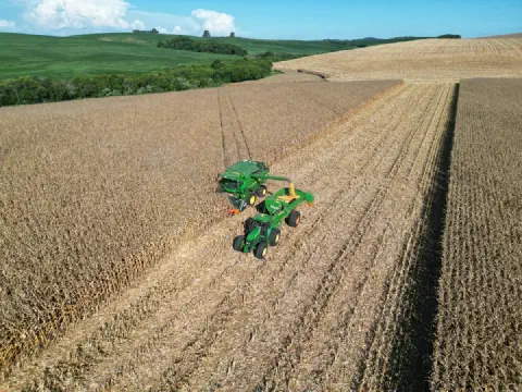 Vista aérea de uma colheitadeira John Deere descarregando milho em um graneleiro puxado por trator, em lavoura de relevo ondulado.