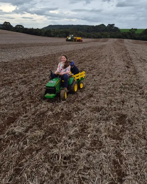 Crianças em um mini trator verde com carreta, em um campo preparado para o plantio, com um trator e uma semeadora ao fundo.