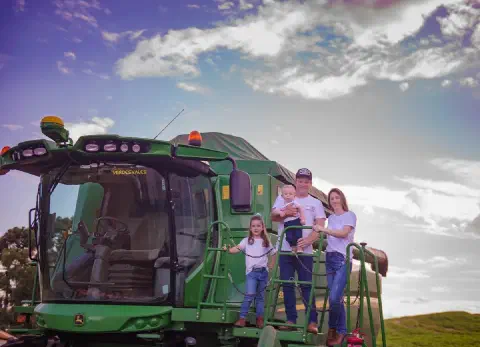 Família em pé na escada de uma colheitadeira John Deere, com céu azul e nuvens ao fundo.