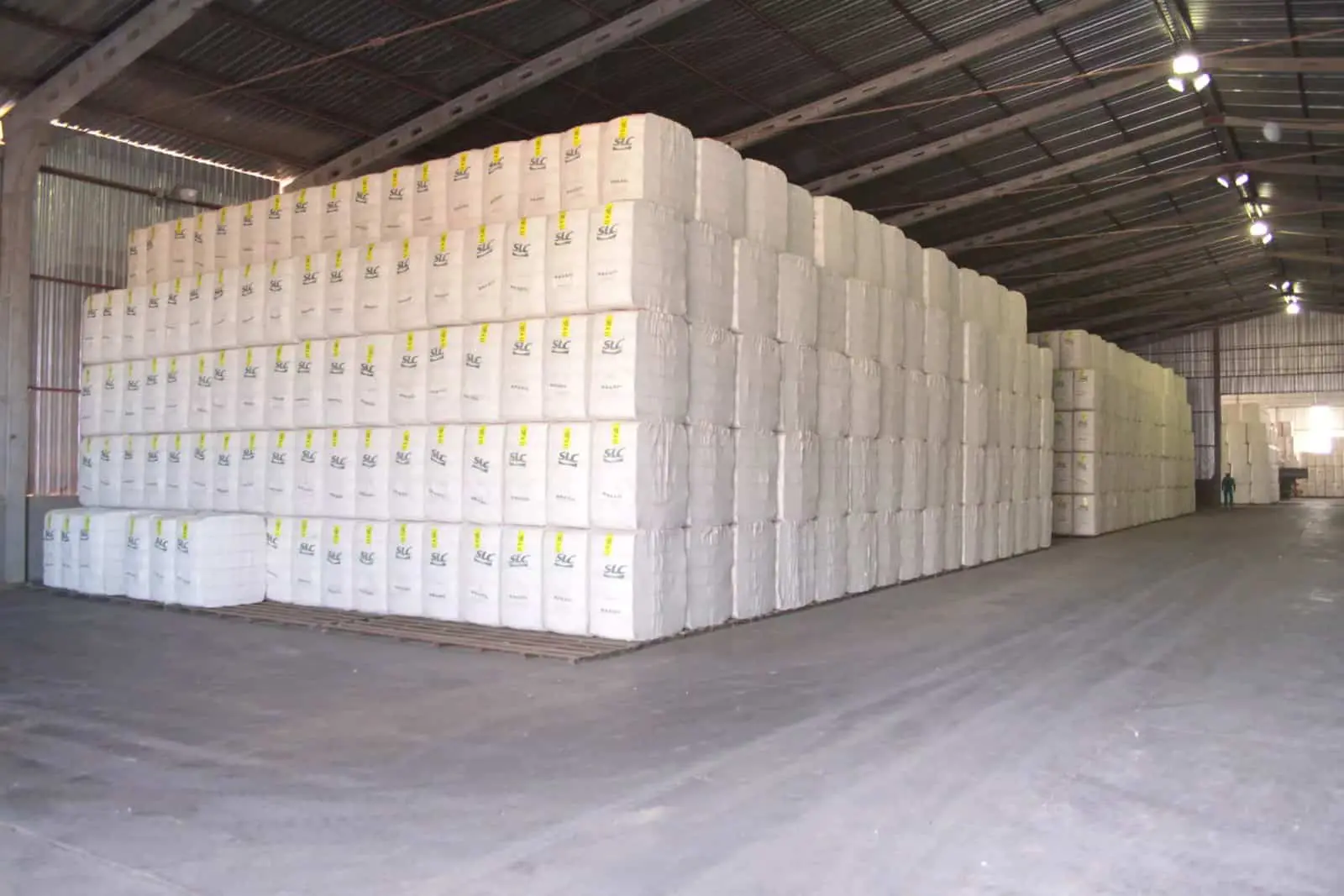 o interior de um amplo galpão de armazenamento agrícola, repleto de gigantescas pilhas de fardos de algodão.