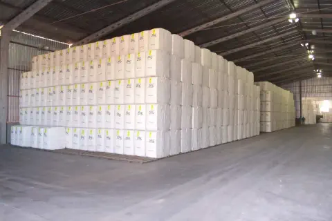 o interior de um amplo galpão de armazenamento agrícola, repleto de gigantescas pilhas de fardos de algodão.