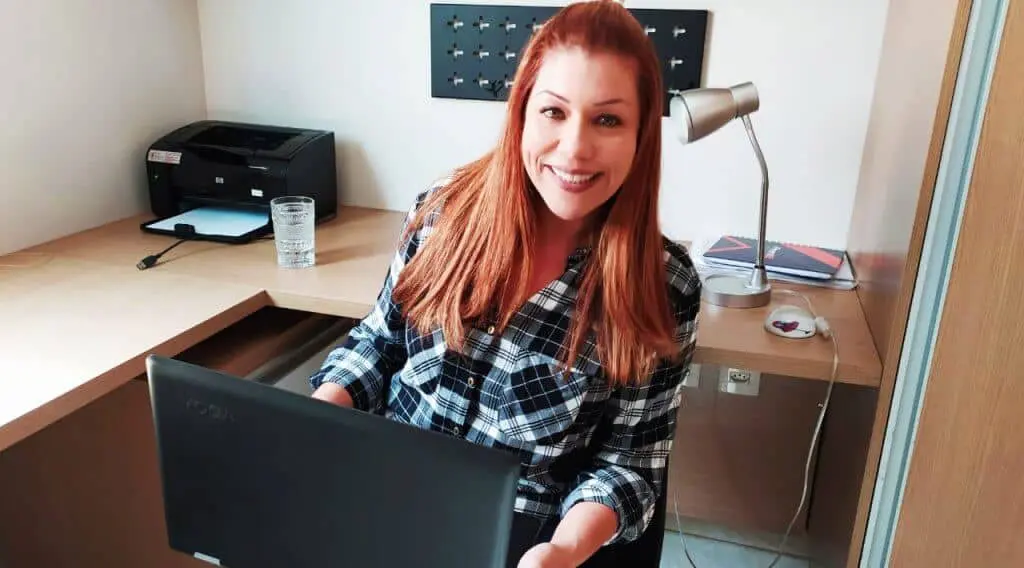 Gestão de fazenda simplificada com software agrícola | Aegro uma mulher sorridente, com cabelos ruivos e vestindo uma camisa xadrez, sentada em seu escritório. Ela segura