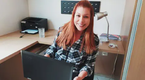 uma mulher sorridente, com cabelos ruivos e vestindo uma camisa xadrez, sentada em seu escritório. Ela segura