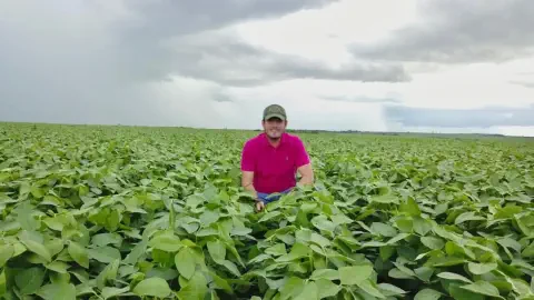 produtor rural sorridente, posicionado no centro de uma vasta e saudável lavoura de soja. Ele veste uma cam