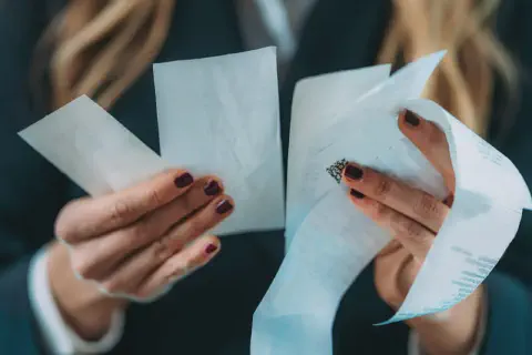 imagem mostra um close-up das mãos de uma mulher, com unhas pintadas em tom escuro, segurando e analisando vários recibos e