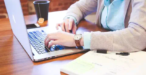uma pessoa sentada em uma mesa de madeira, digitando em um laptop prateado. O foco está nas mãos da pessoa sob