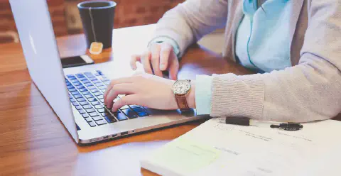 uma pessoa sentada em uma mesa de madeira, digitando em um laptop prateado. O foco está nas mãos da pessoa sob