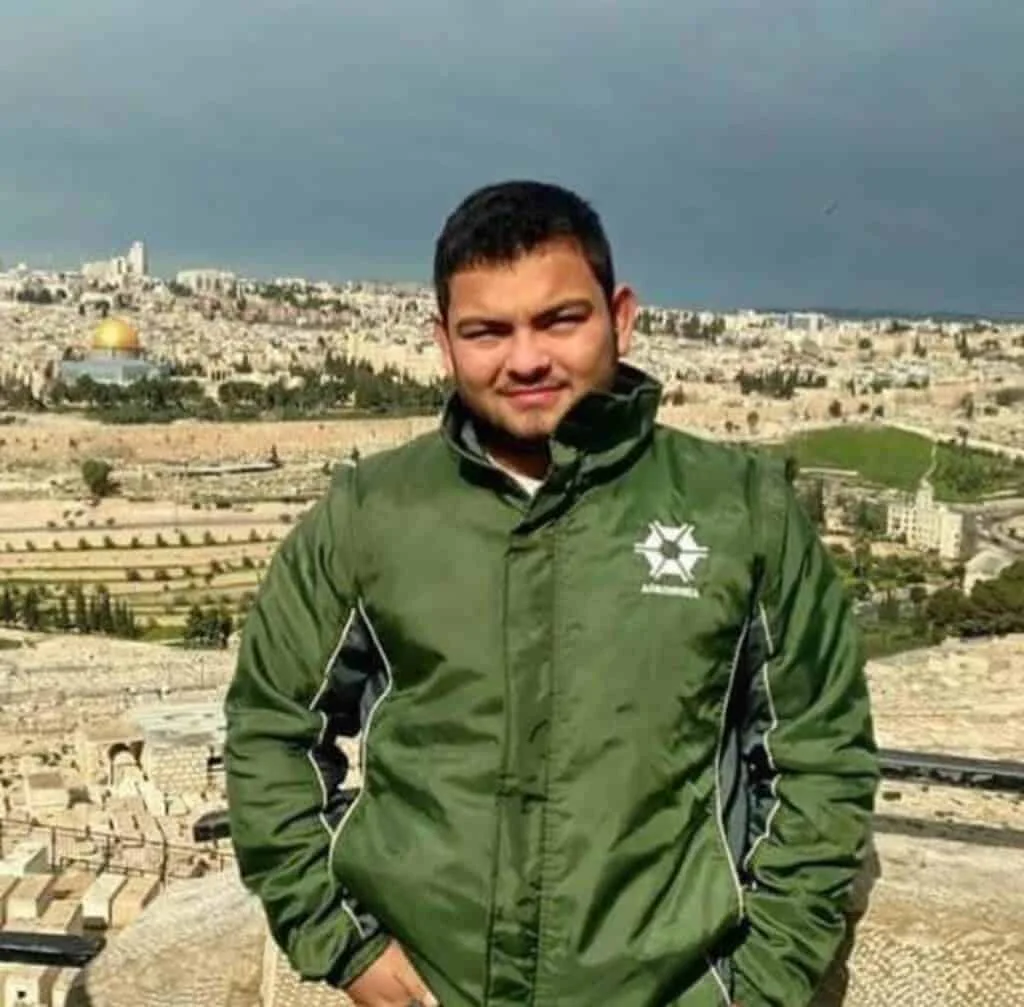 Homem em viagem com vista panorâmica de Jerusalém jovem em primeiro plano, posando para uma foto em um ponto de observação elevado. Ele veste uma jaqueta v