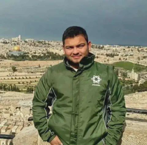 jovem em primeiro plano, posando para uma foto em um ponto de observação elevado. Ele veste uma jaqueta v