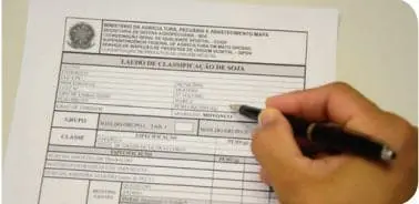 close-up da mão de uma pessoa preenchendo um documento oficial com uma caneta preta. O formulário é um ‘Lau