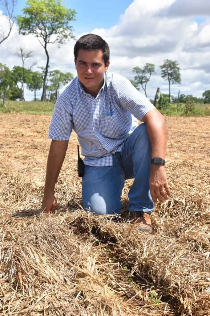 homem, provavelmente um agrônomo ou produtor rural, ajoelhado em um campo agrícola sob um céu azul com nu
