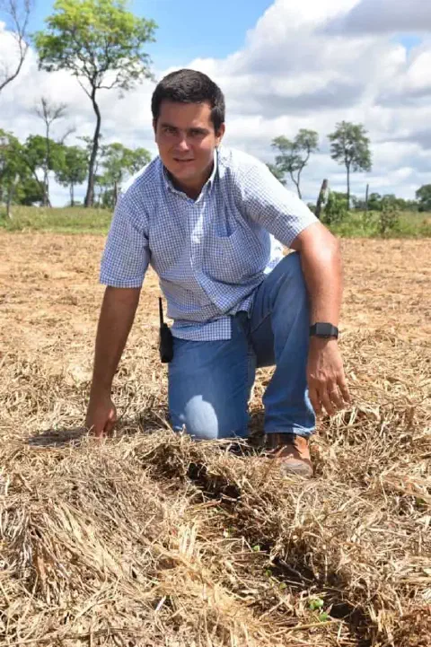 homem, provavelmente um agrônomo ou produtor rural, ajoelhado em um campo agrícola sob um céu azul com nu