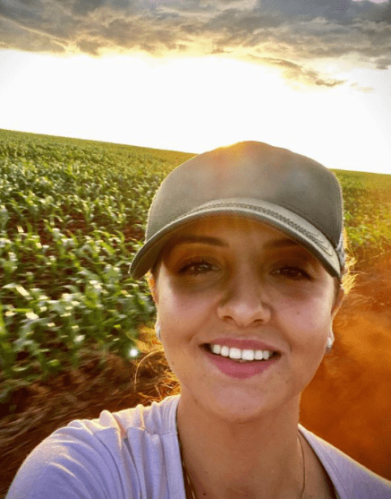 Mulher no Agro: Gestão e Sucesso na Lavoura Moderna selfie tirada por uma mulher jovem e sorridente, que usa um boné. Ela está em uma grande lavoura, possivelment