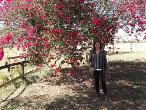 mulher sorridente em um ambiente rural ensolarado, posicionada em frente a uma exuberante árvore de prim