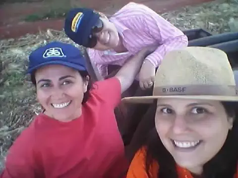 selfie casual de três mulheres sorrindo em um ambiente rural, provavelmente uma fazenda ou campo agrícola. O f