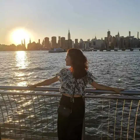 mulher de costas, apoiada em uma grade, contemplando a silhueta de uma grande metrópole ao pôr do sol. A