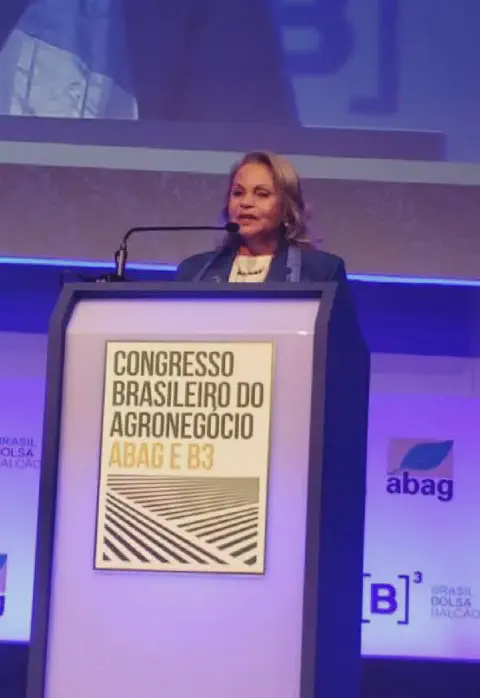 mulher discursando em um púlpito durante um evento formal. Ela está vestida profissionalmente com um bla