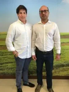 dois homens, em pé, lado a lado, posando para uma foto em um ambiente interno, provavelmente um escritório. Am