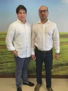 dois homens, em pé, lado a lado, posando para uma foto em um ambiente interno, provavelmente um escritório. Am