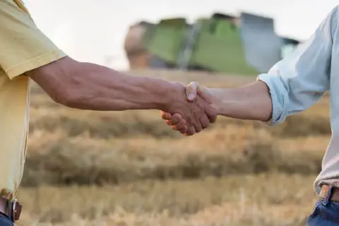 A imagem captura em primeiro plano o aperto de mãos firme entre dois homens em um ambiente rural. O cenário de fundo, desfoca