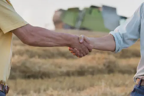 A imagem captura em primeiro plano o aperto de mãos firme entre dois homens em um ambiente rural. O cenário de fundo, desfoca