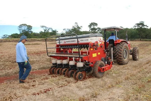 operação de plantio mecanizado em uma área rural. Em primeiro plano, um trator vermelho de grande porte
