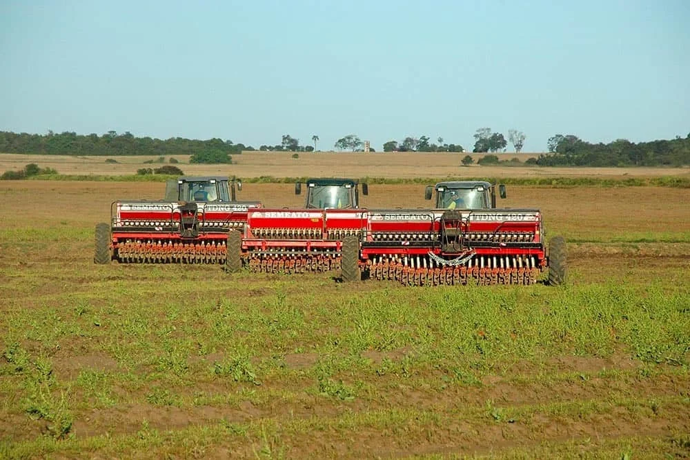 três tratores alinhados em um vasto campo agrícola, cada um acoplado a uma grande semeadora vermelha da marca