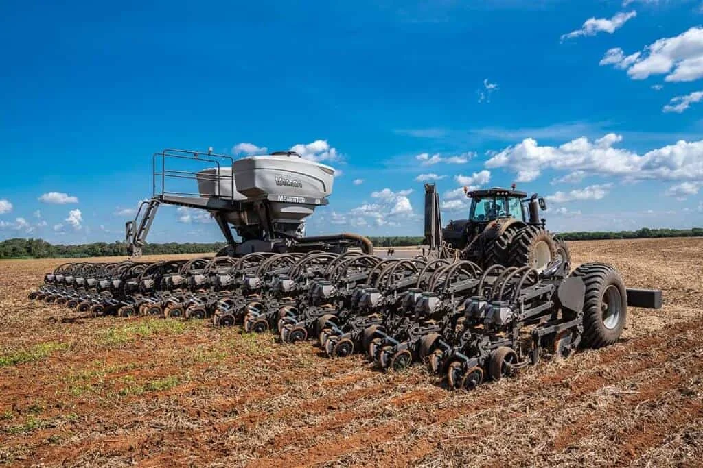 uma operação de plantio em larga escala em um dia ensolarado. Em primeiro plano, um trator moderno e robusto e