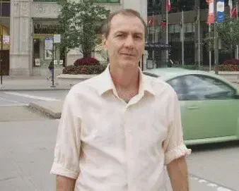 o retrato de um homem de meia-idade, posicionado em um ambiente urbano. Ele veste uma camisa social de cor cla