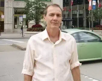 o retrato de um homem de meia-idade, posicionado em um ambiente urbano. Ele veste uma camisa social de cor cla