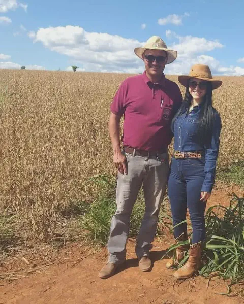 casal, um homem e uma mulher, em pé na beira de uma vasta lavoura de soja. Ambos vestem trajes caracterís