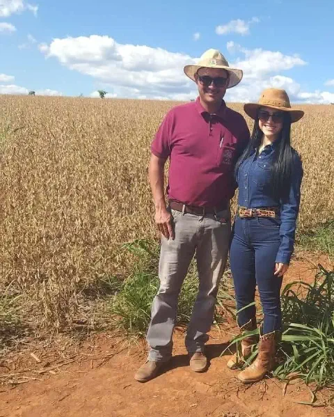 casal, um homem e uma mulher, em pé na beira de uma vasta lavoura de soja. Ambos vestem trajes caracterís