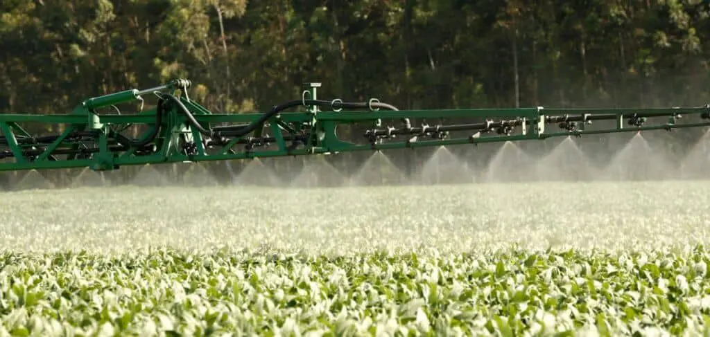 uma operação de pulverização agrícola em uma extensa lavoura, provavelmente de soja, em um dia ensolarado. Em