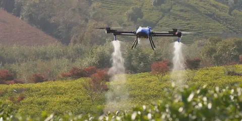 drone agrícola profissional, de cor azul e preta, em pleno voo sobre uma extensa plantação. O equipamento e