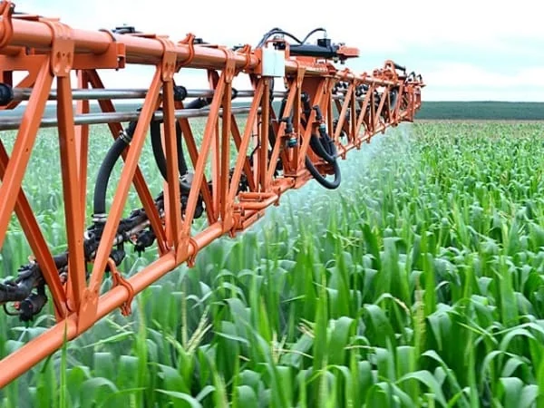 primeiro plano a longa barra de um pulverizador agrícola de cor laranja, em plena operação sobre uma extens