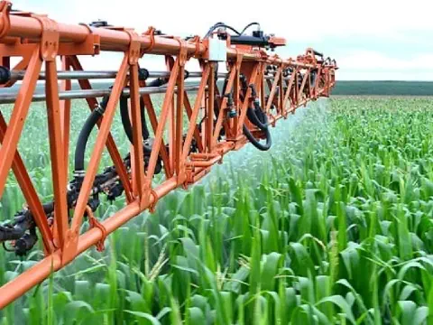 primeiro plano a longa barra de um pulverizador agrícola de cor laranja, em plena operação sobre uma extens