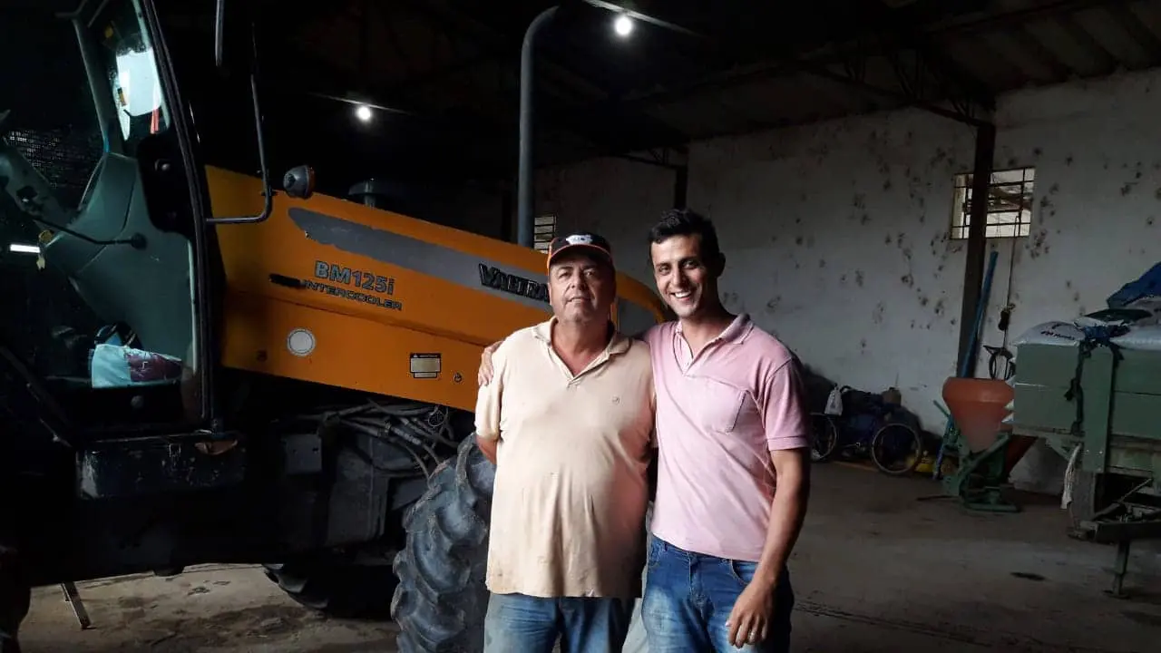 Sucessão Familiar no Agronegócio: Pai e Filho na Fazenda dois homens, aparentemente pai e filho, posando juntos dentro de um galpão agrícola. O ambiente é rústico e