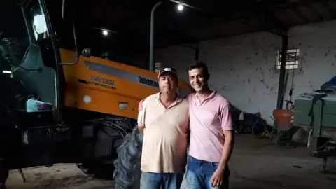 dois homens, aparentemente pai e filho, posando juntos dentro de um galpão agrícola. O ambiente é rústico e