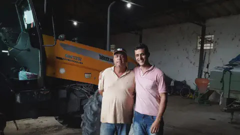 dois homens, aparentemente pai e filho, posando juntos dentro de um galpão agrícola. O ambiente é rústico e