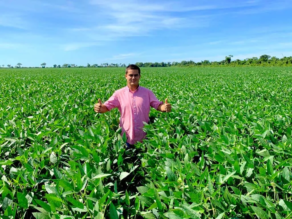 agricultor de pé, no meio de uma vasta e próspera lavoura de soja. O homem, vestindo uma camisa rosa, sorri