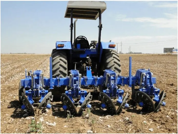 a traseira de um trator azul de médio porte, parado em uma lavoura sob um céu claro e ensolarado. Acoplado ao